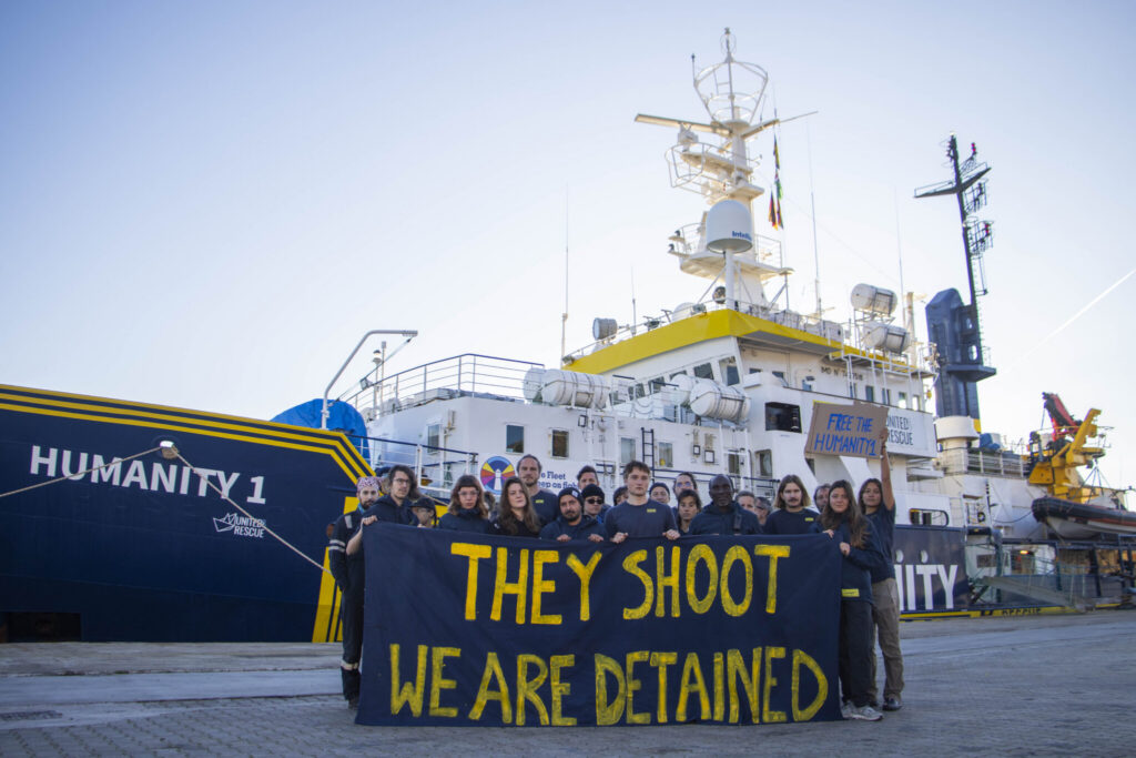 Eine Gruppe von etwa zwanzig Menschen steht auf einem Hafen vor dem Rettungsschiff „HUMANITY 1“. Sie halten ein großes dunkelblaues Banner mit gelber Schrift, auf dem steht: „THEY SHOOT WE ARE DETAINED“. Eine Person hält zusätzlich ein Schild mit der Aufschrift „FREE THE HUMANITY 1“. Das Schiff im Hintergrund trägt die Logos „HUMANITY 1“ und „UNITED4RESCUE“.