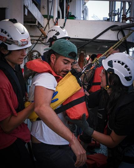 Ein Mann mit umgedrehter grüner Cap und weißem T-Shirt wird von zwei Helferinnen der Sea Punks versorgt; ihm wird eine orange-gelbe Rettungsweste angelegt. Im Hintergrund Seile, Karabiner und weitere Crew an der Schiffsreling. Übung zur Patientenversorgung.