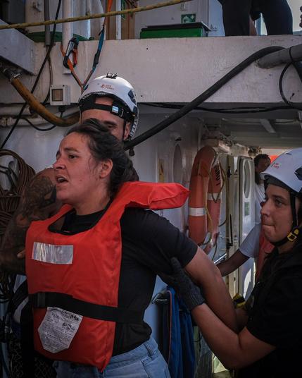 Eine Frau in oranger Rettungsweste mit geöffnetem Mund (sie ruft oder spricht) wird von hinten von einem Crewmitglied der Sea Punks gestützt. Im Hintergrund ein orange-weißer Rettungsring an der weißen Schiffswand. Trainingsszenario.