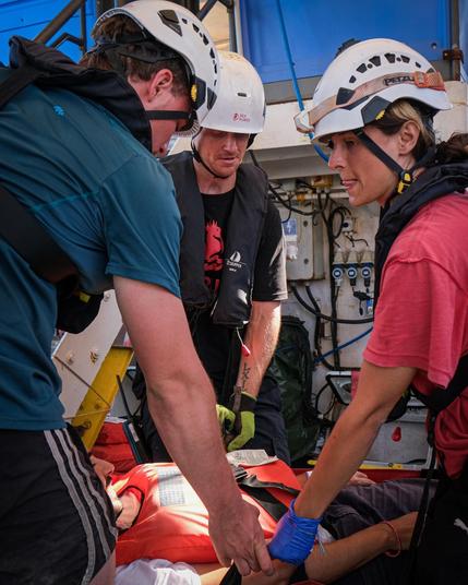 Mehrere Crewmitglieder der Sea Punks mit Helmen knien und beugen sich über eine Person, die auf einem roten Spineboard (Rettungstrage) liegt, und legen Hand an. Übung zur Erstversorgung an Deck.