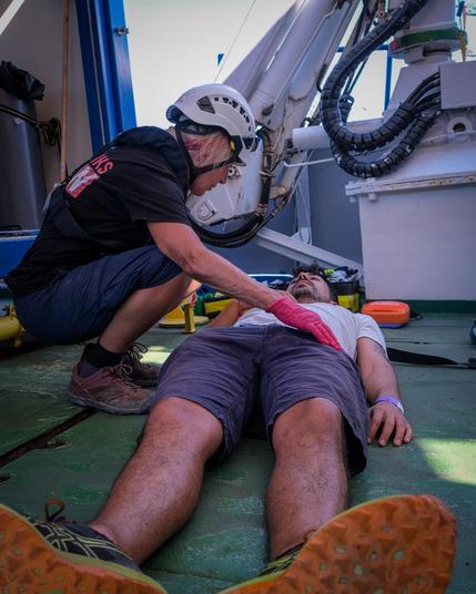 Eine Helferin der Sea Punks mit weißem Helm kniet über einem Mann, der als Patient in Shorts und T-Shirt auf dem grünen Schiffsdeck liegt, und untersucht ihn. Im Hintergrund Bordtechnik und Schläuche. Übung zur Patientenbeurteilung.