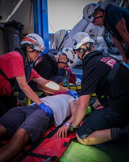 Eine Gruppe von Crewmitgliedern der Sea Punks mit Helmen versorgt mit blauen Einmalhandschuhen einen Mann, der auf einem roten Spineboard auf dem grünen Deck liegt. Im Hintergrund Schiffsaufbauten und Seile. Massenanfall-Training.