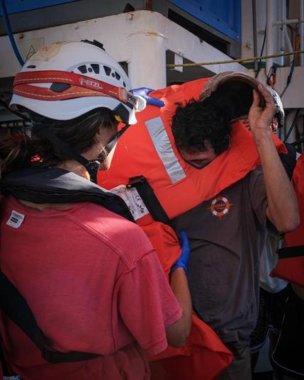 Eine Helferin der Sea Punks mit weißem Helm, von hinten gesehen, stützt einen Mann in oranger Rettungsweste, der sich erschöpft nach vorne beugt. Enge Szene zwischen Schiffsaufbauten.
