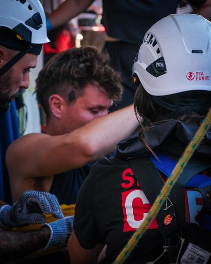 Nahaufnahme: Ein bärtiger Helfer der Sea Punks mit Helm und ein junger Mann (Patientendarsteller) blicken konzentriert nach unten; rechts ein Crewmitglied mit Helm und schwarzem Shirt, von hinten. Dichtes Gedränge während der Übung.